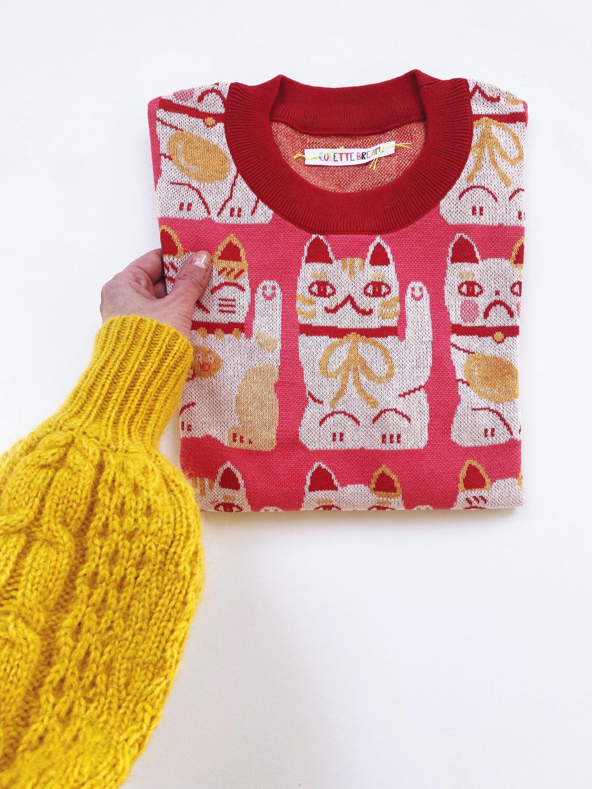 Folded pink and red sweater with maneki neko cat pattern held by a person wearing a yellow sweater.