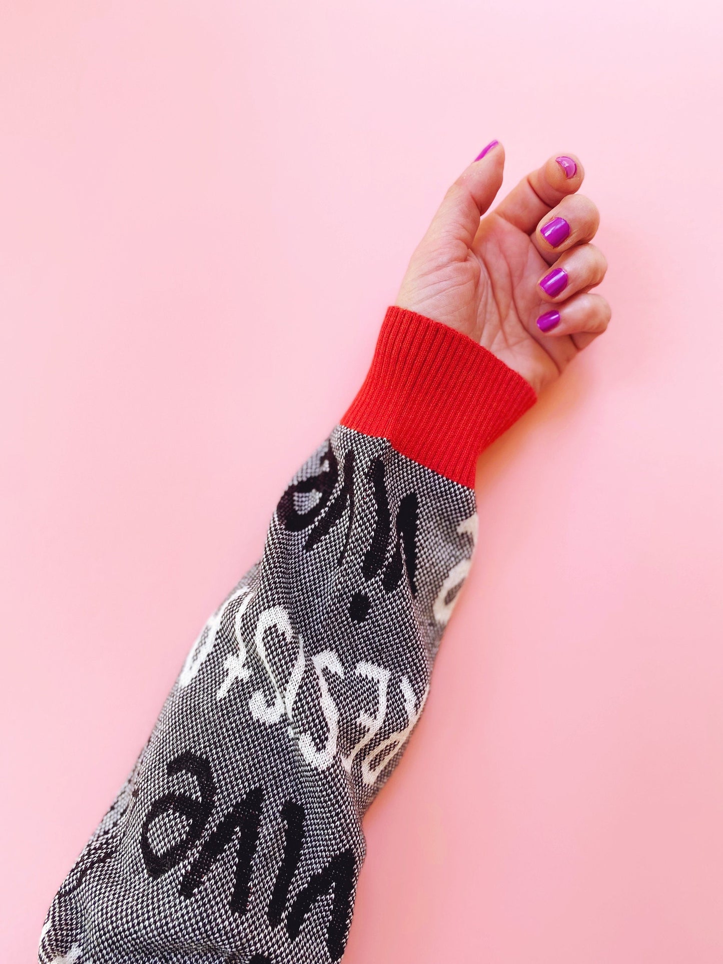 Hand wearing a black and white patterned sweater with red cuff against a pink background