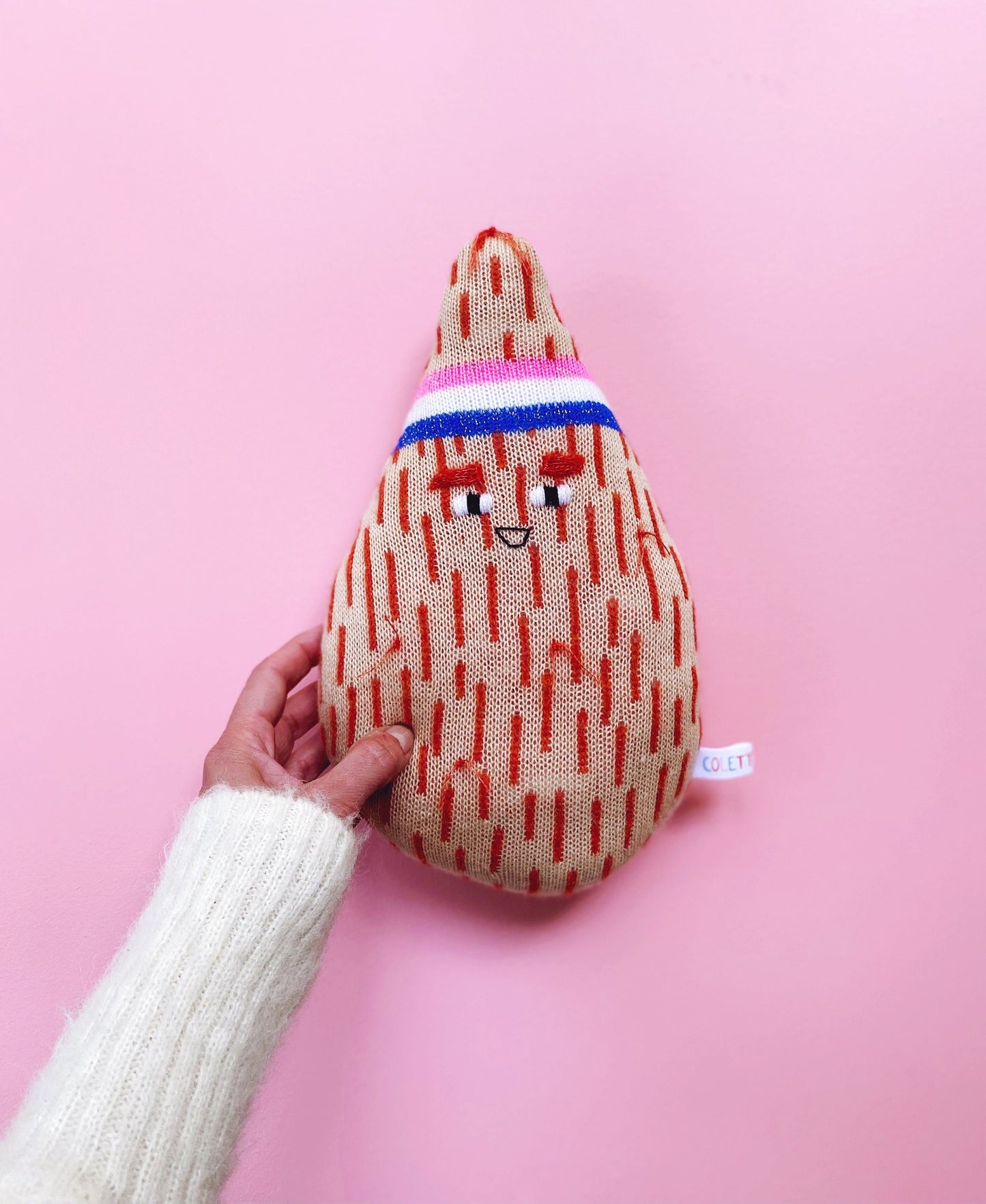Hand holding a knitted plush coconut toy with a patterned design against a pink background