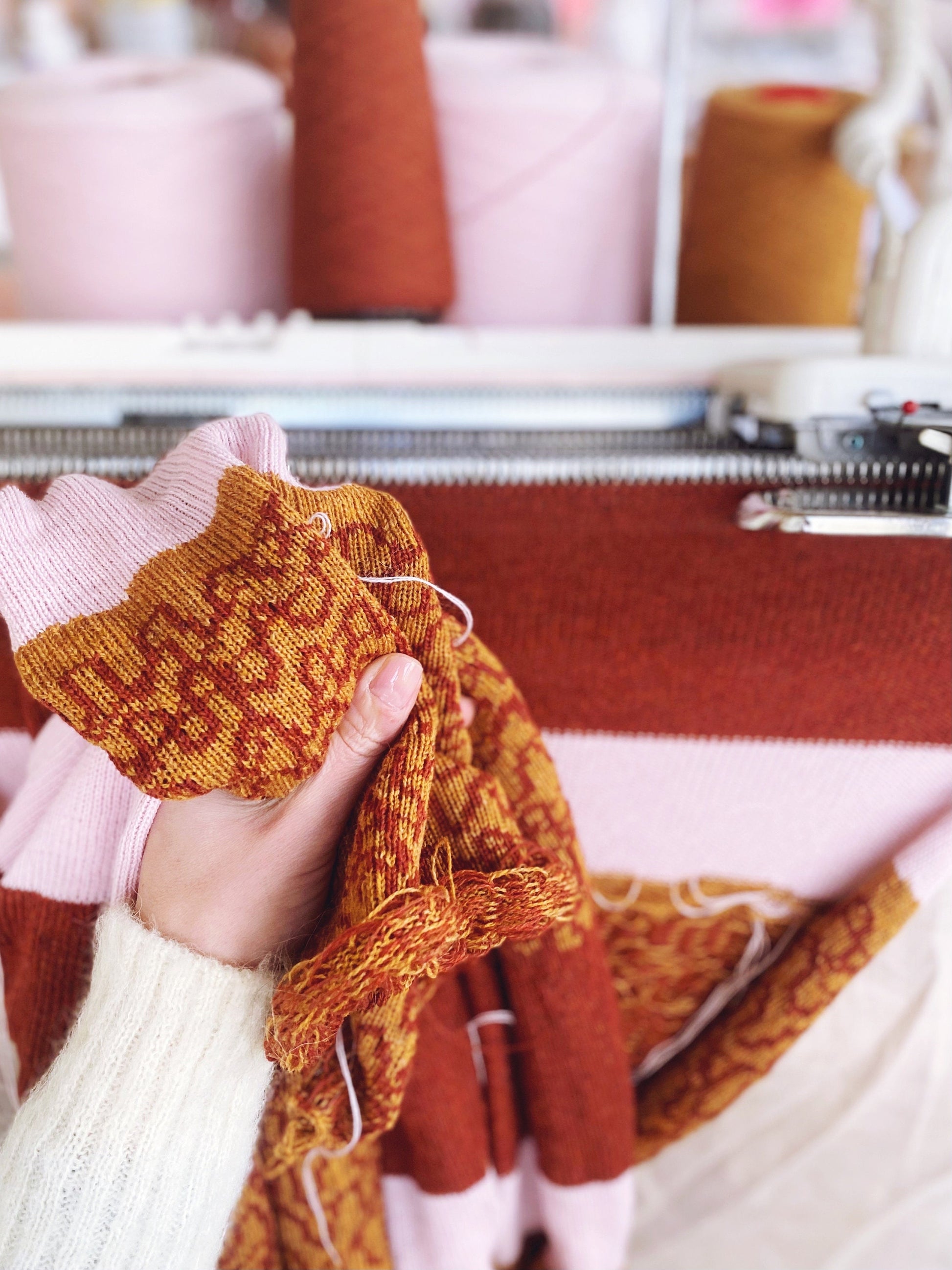 Hand holding a knitted pattern in front of a knitting machine with spools of yarn in the background.