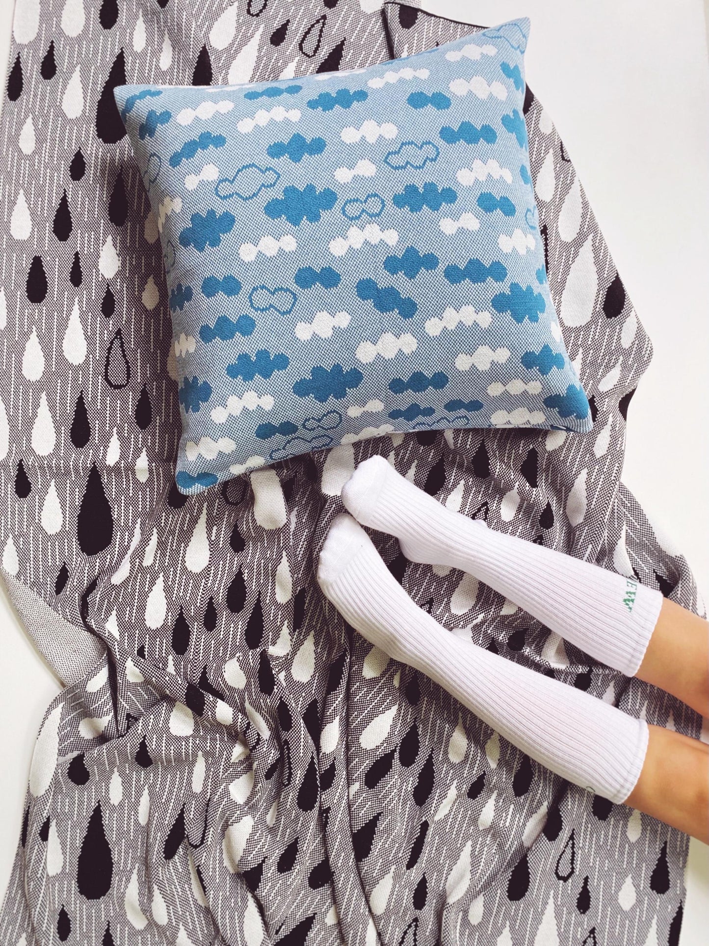 Blue pillow with cloud pattern on a knitted blanket with raindrop pattern, next to white socks.