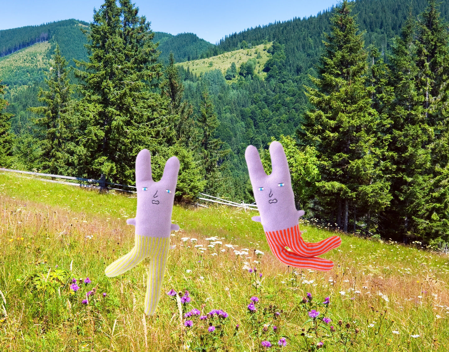 Two cartoonish rabbit plush toys in a field with mountains and trees and wildflowers in the background