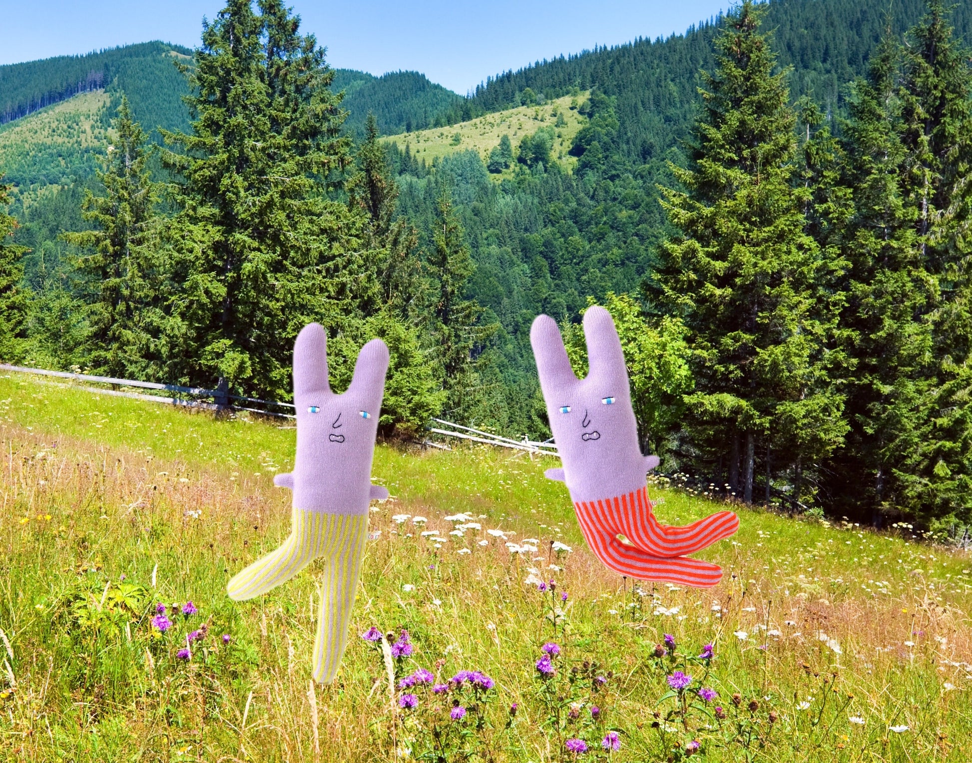 Two cartoonish rabbit plush toys in a field with mountains and trees and wildflowers in the background