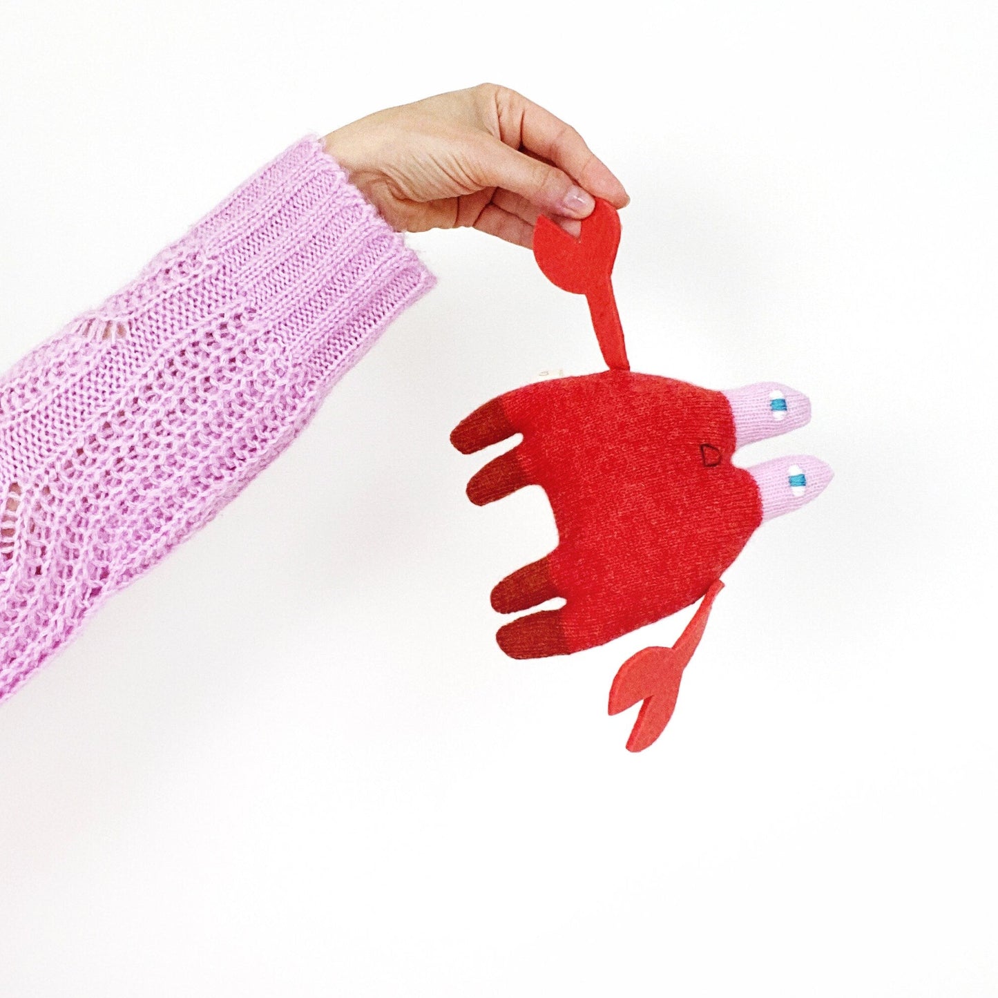 Red knitted lambswool crab-shaped toy held by a hand against a white background