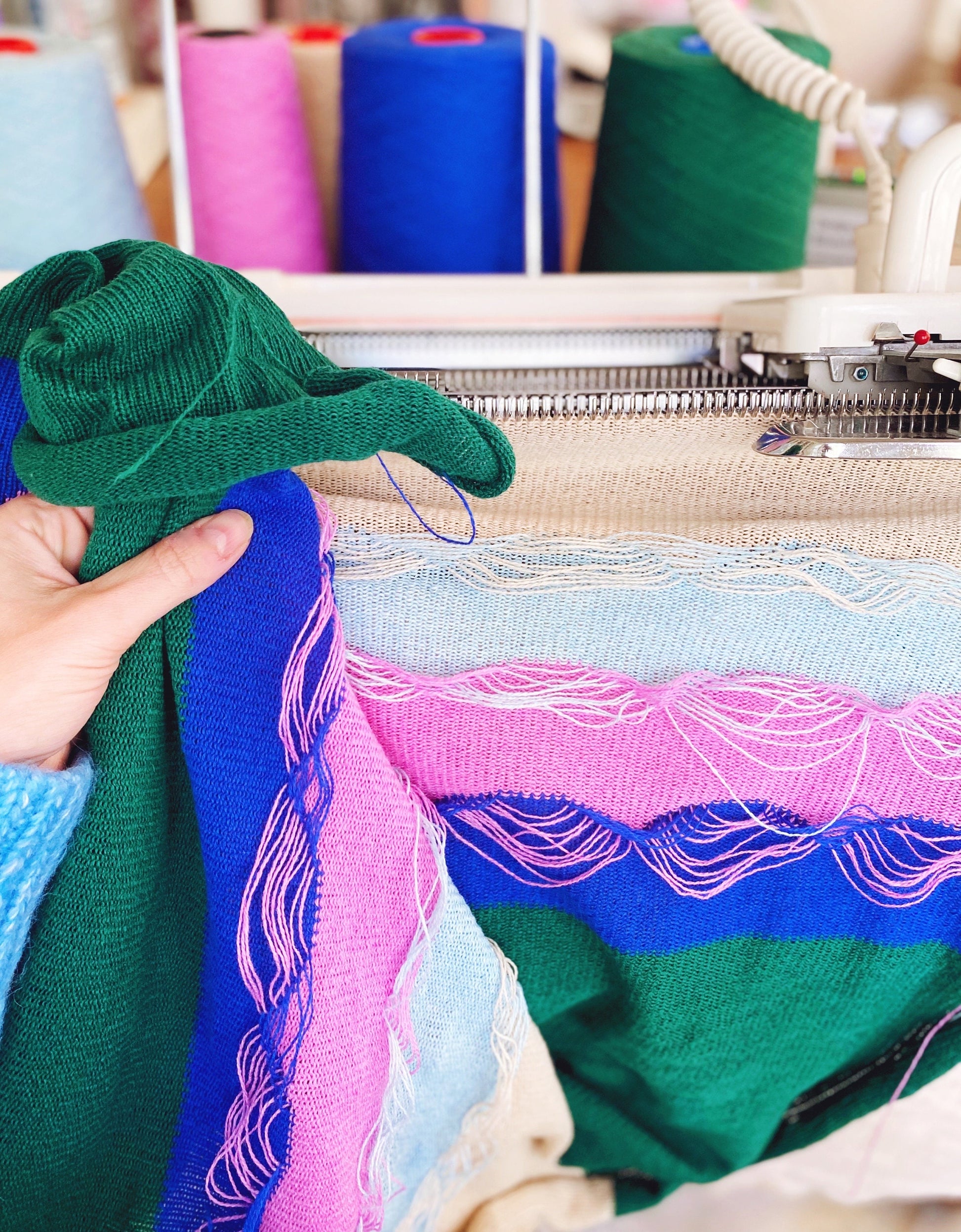 Colorful textured fabric being held up next to a knitting machine.