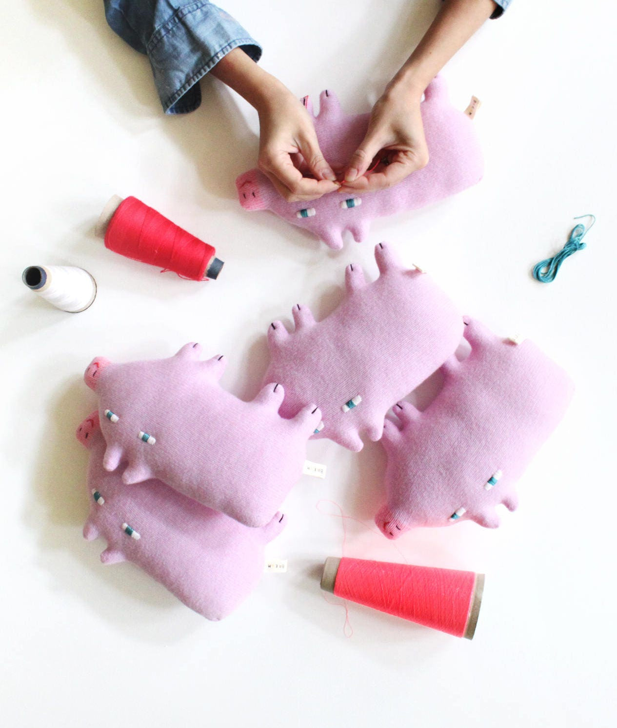 Pink knitted piglet plush toys being worked on with sewing materials on a white surface