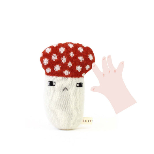 Knitted mushroom baby rattle toy with a red cap and white spots, next to a pink hand on a white background.