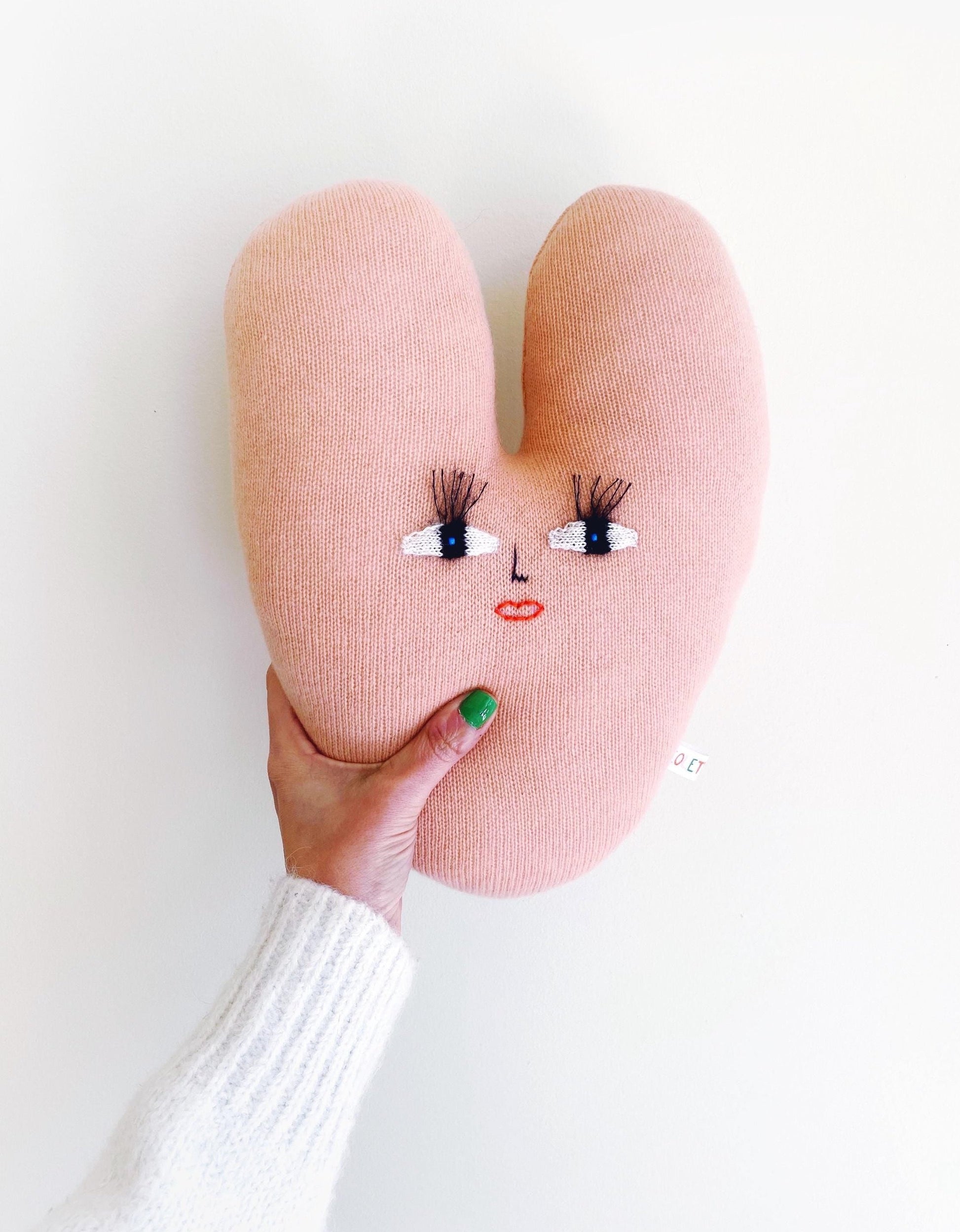 Heart-shaped knitted pink pillow with face details held by a hand on a white background