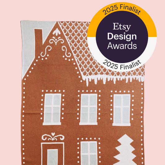 Knitted blanket with gingerbread house design and Etsy Design Awards 2025 Finalist badge on a pink background