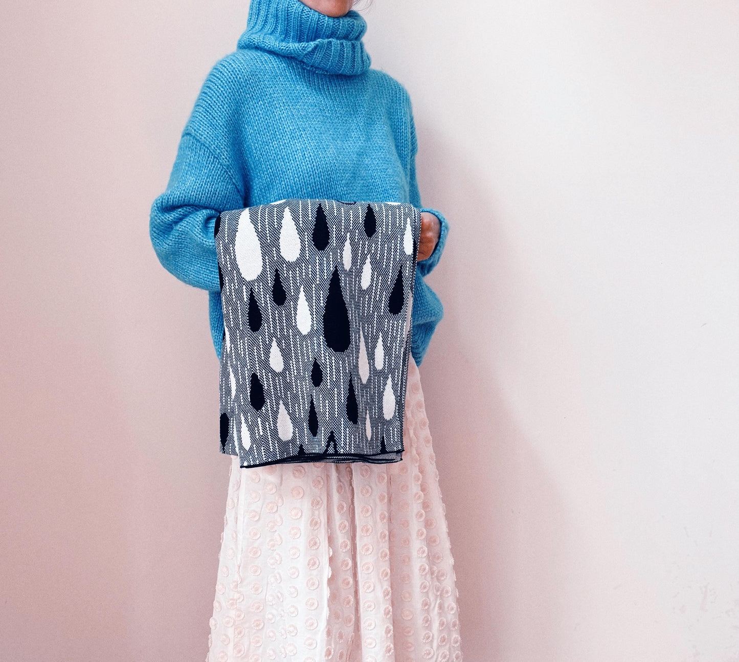 Person wearing a blue sweater holding a gray knitted blanket with black and white raindrop pattern against a light pink backgroundted 
