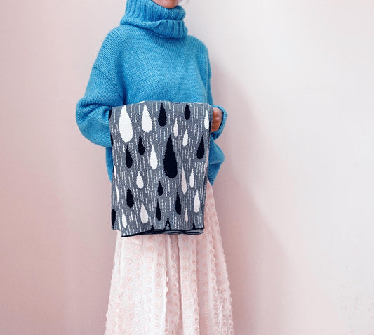 Person wearing a blue sweater holding a gray knitted blanket with black and white raindrop pattern against a light pink backgroundted 
