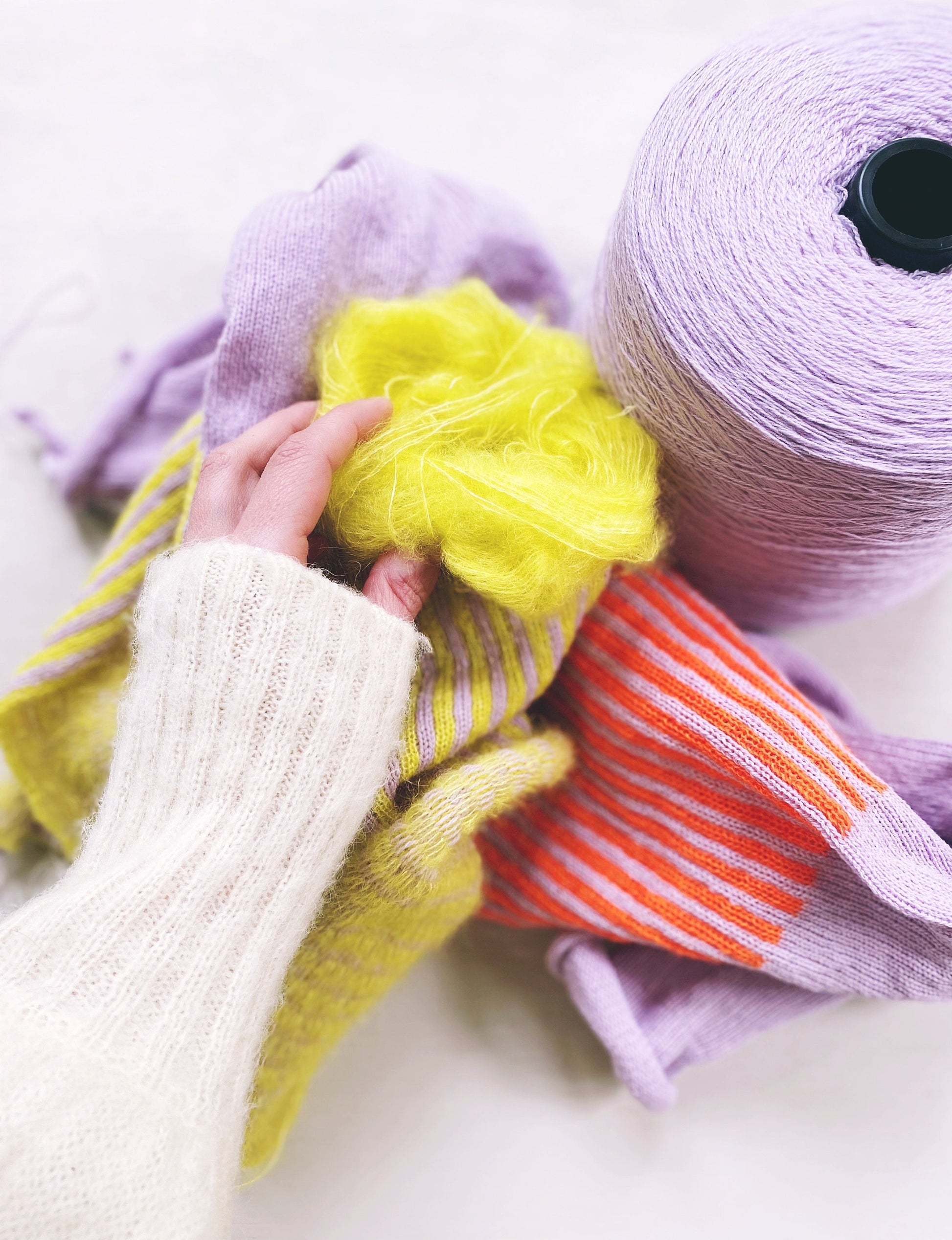 Hand holding a ball of yellow yarn next to spools of purple and orange yarn on a white background