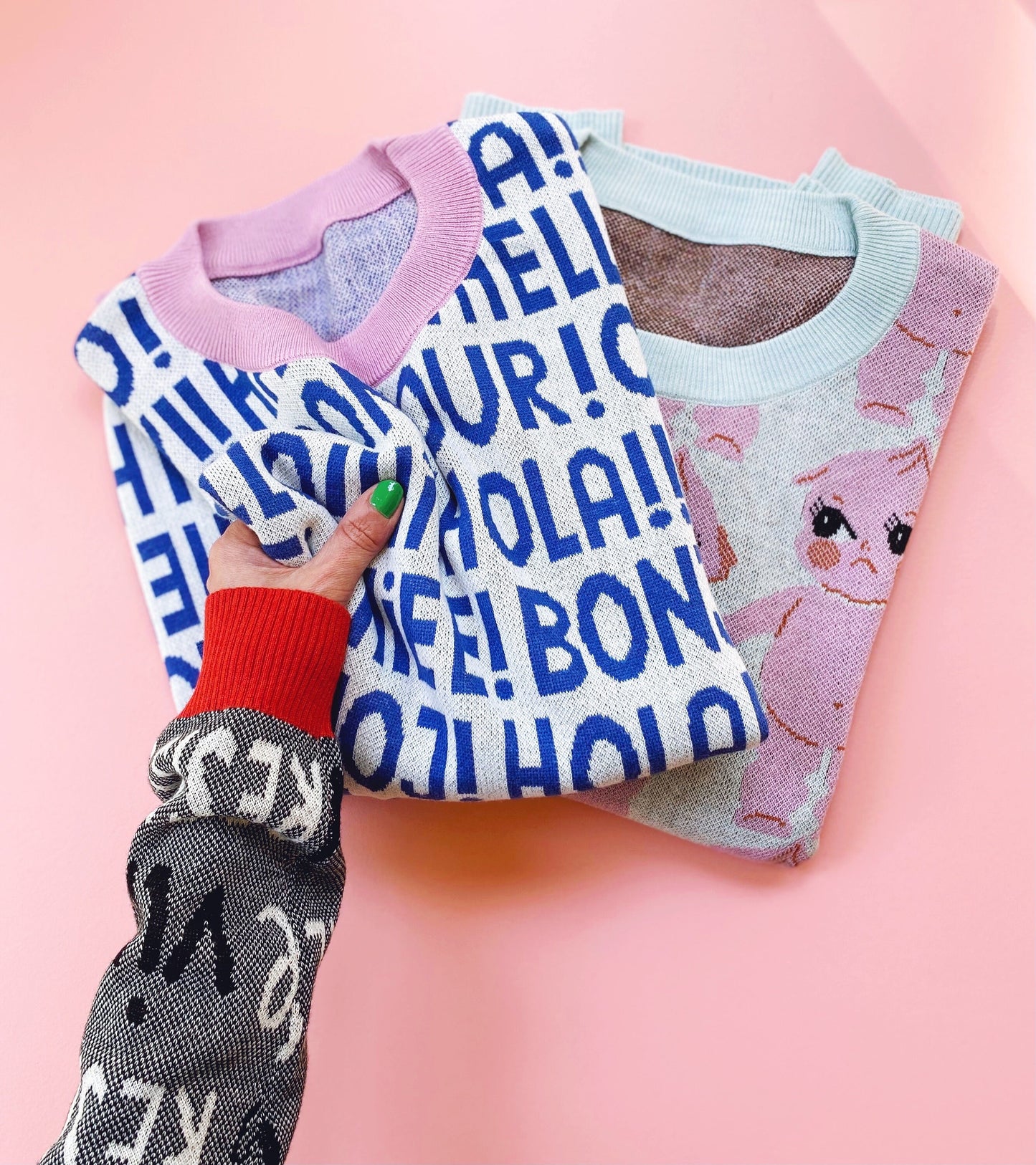 A hand holding two folded sweaters with different designs on a pink background