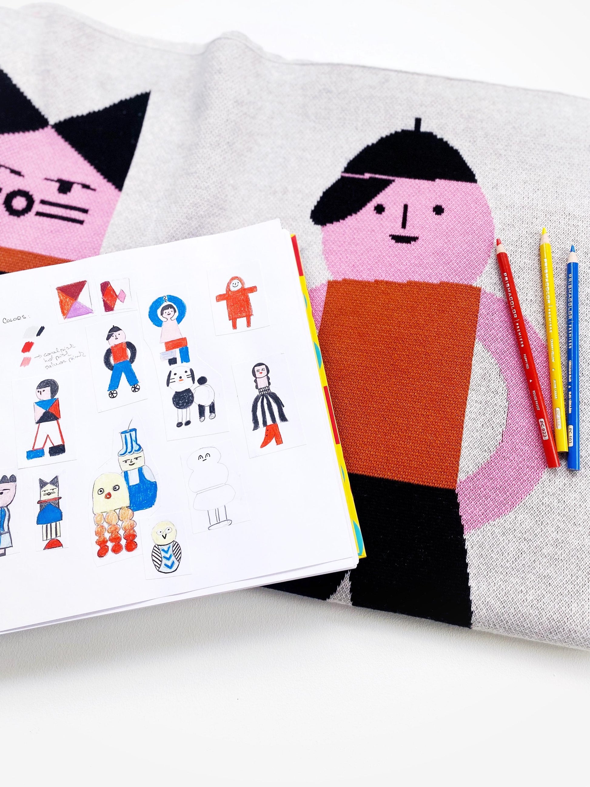 Colorful children's book with illustrations of people, a knit blanket with a cartoon face, and colored pencils on a white background.
