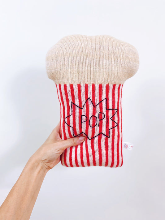 Hand holding a red and white striped popcorn shaped pillow with 'Pop' design against a white background