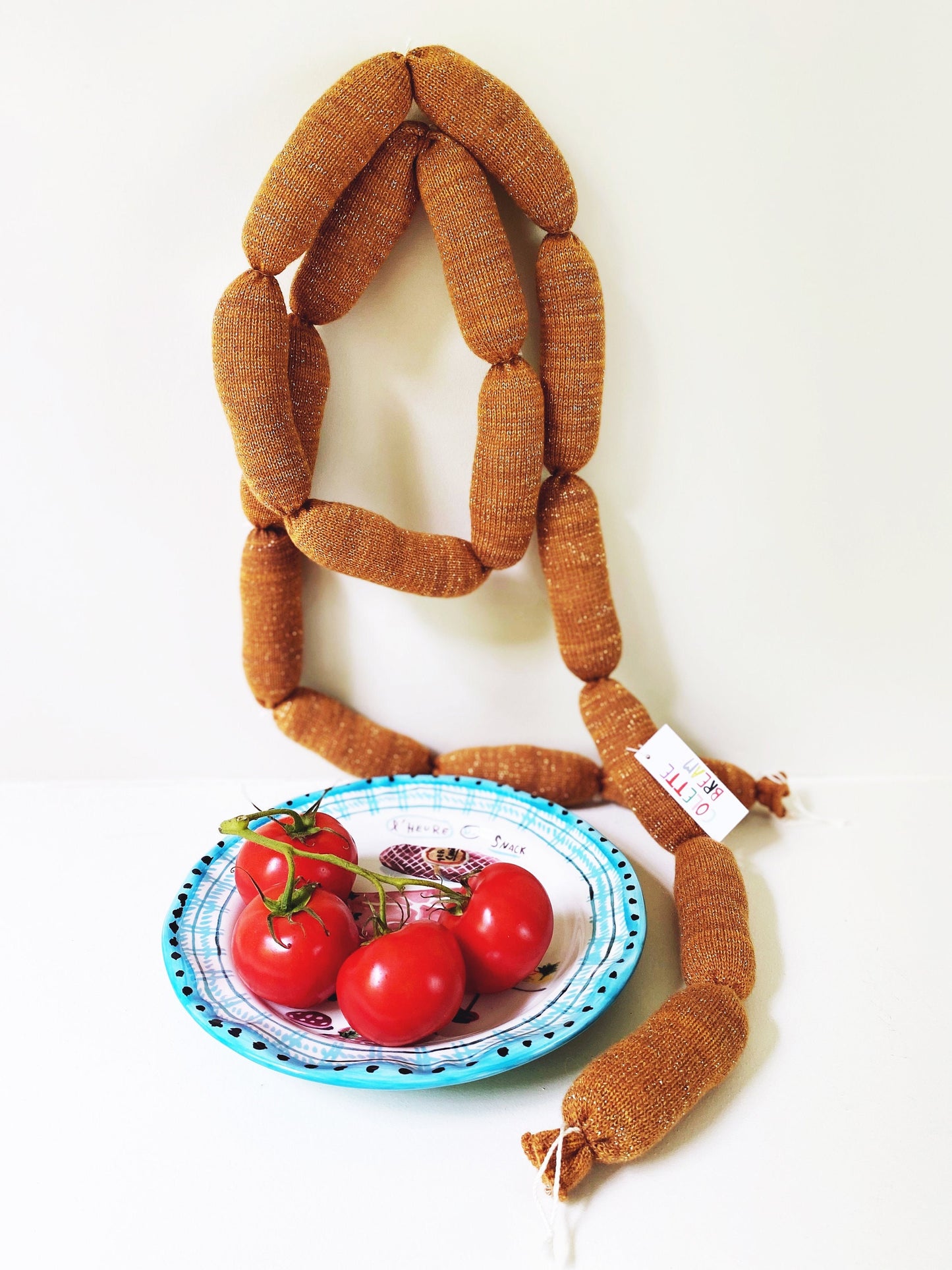 Long brown knitted sausage with a plate of red tomatoes on a white background