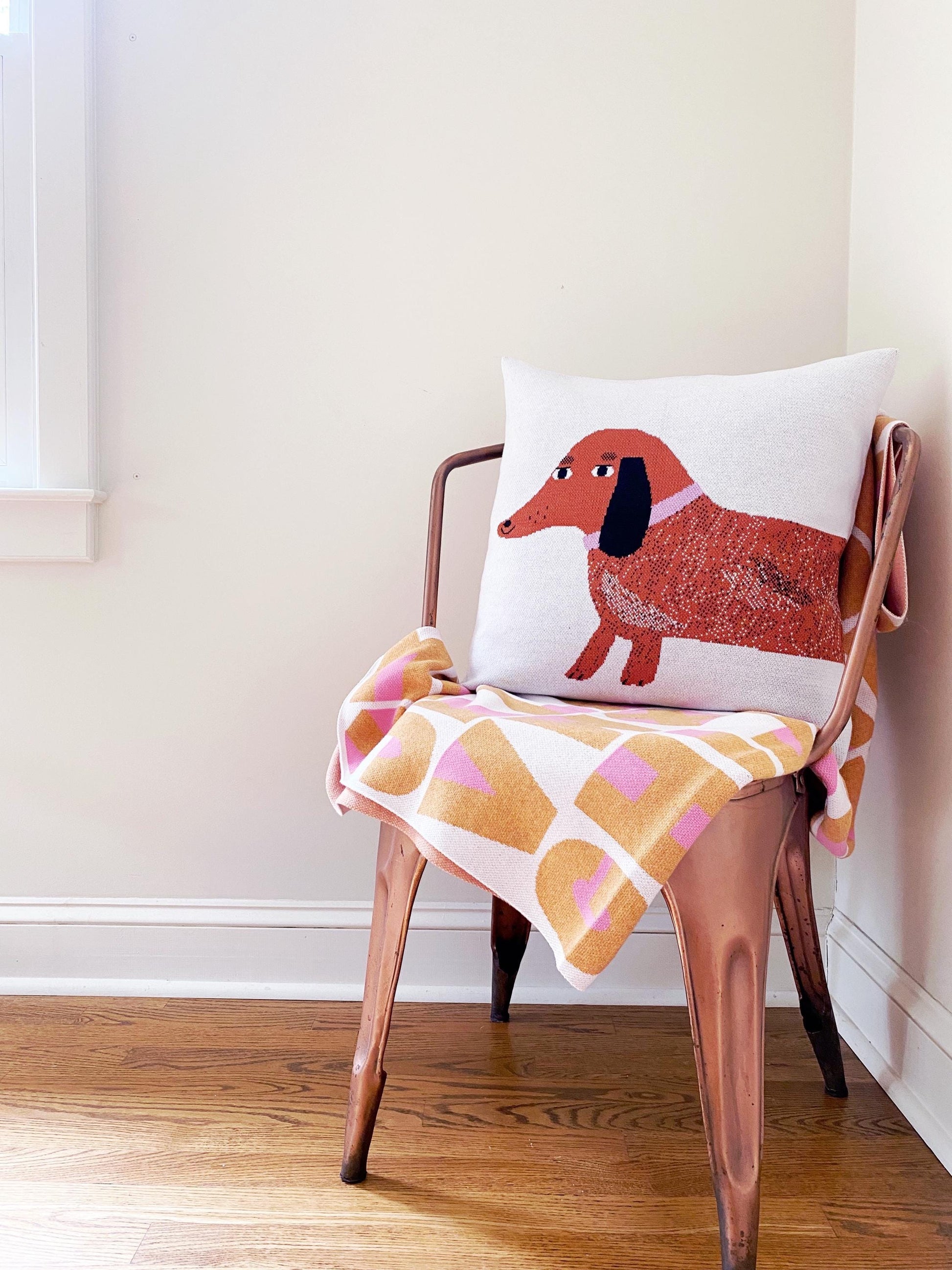 Chair with a pillow featuring a dachshund dog design and a checkered blanket on a wooden floor.