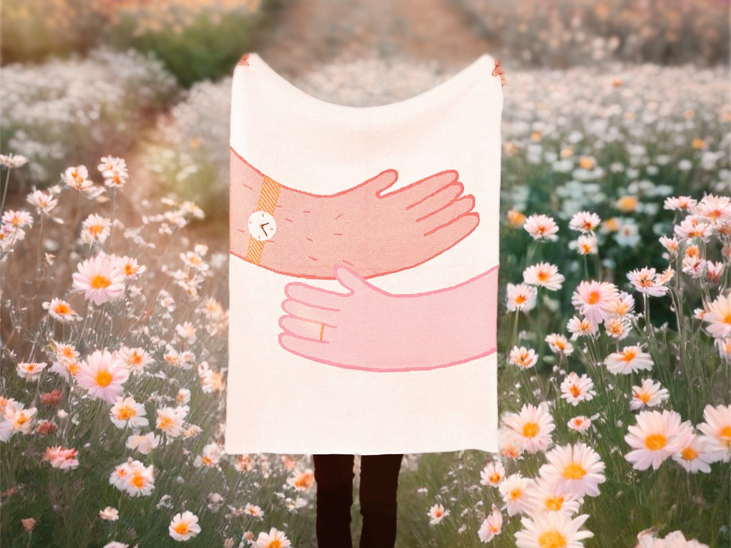 Person holding a knit blanket with a design of two pink hands embracing, standing in a field of flowers.