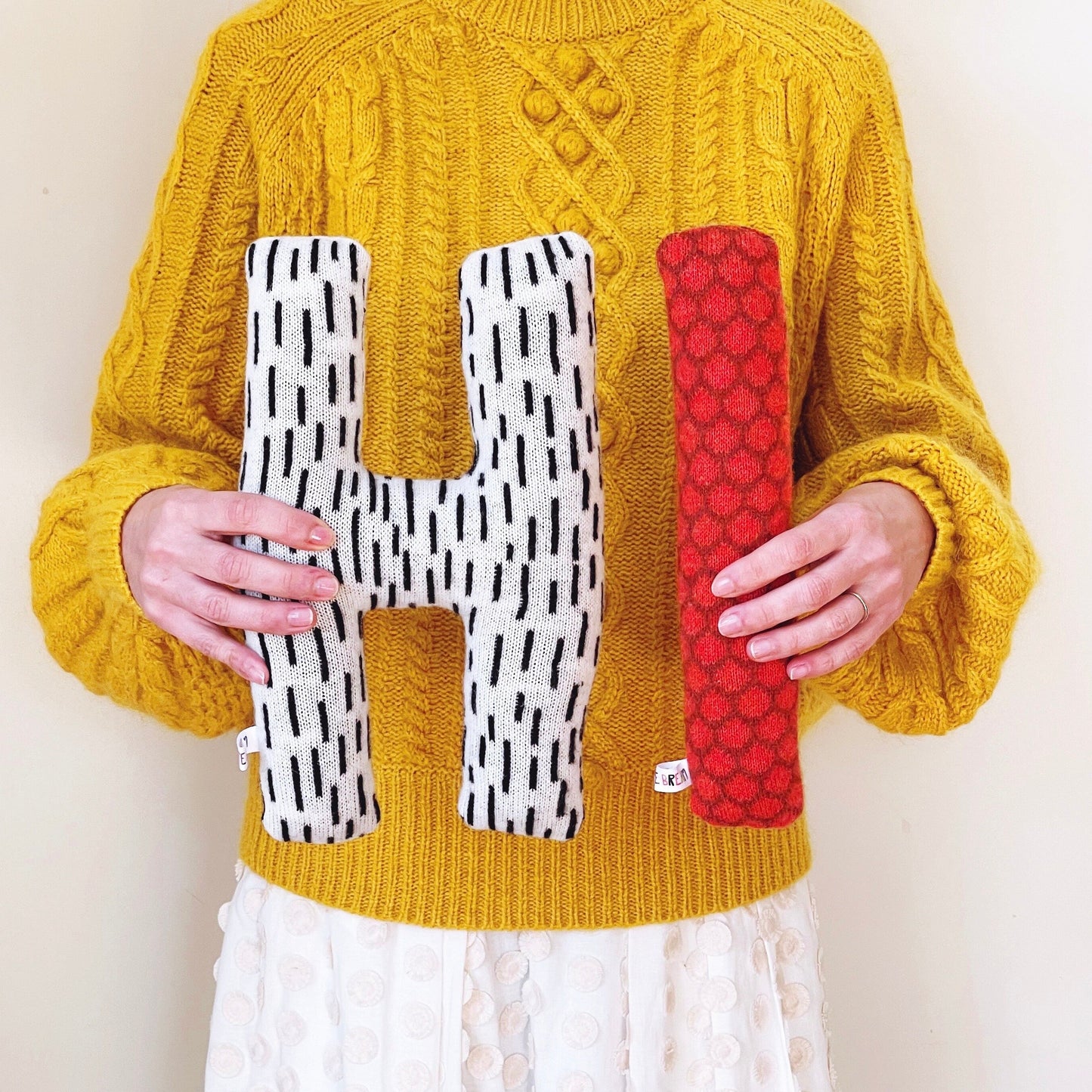 Person wearing a yellow sweater holding two colorful letter shaped pillows 'H' and 'I'.