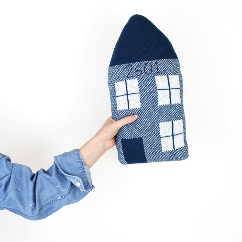 Hand holding a blue knitted house-shaped pillow with white windows on a white background