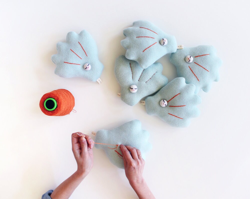 Hands working on a small blue shell shaped plush toy with a ball of yarn on a white background