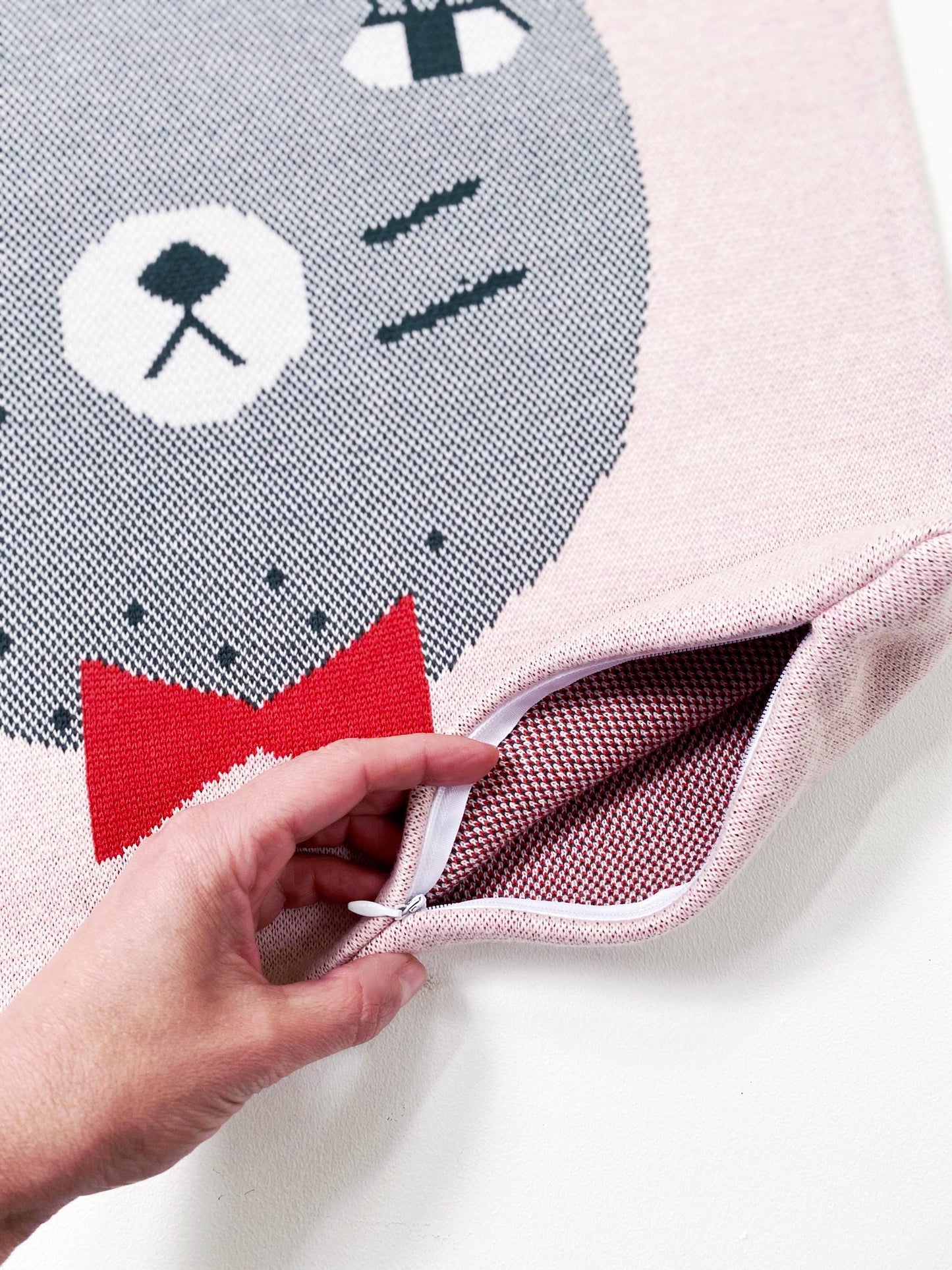 Close-up of a knitted cushion with a cat design and a hand opening a zipper.