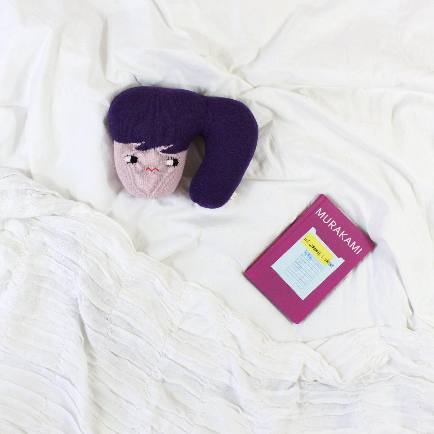 Plush knit toy with purple hair and pink face on a white blanket next to a pink book titled 'Murakami'.
