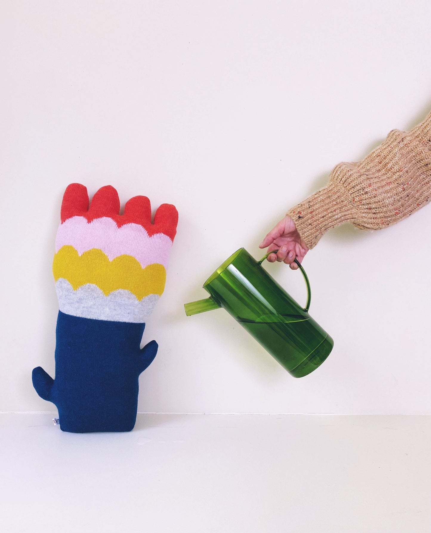 Colorful knitted flower shaped pillow with a hand holding a green watering can on a white background