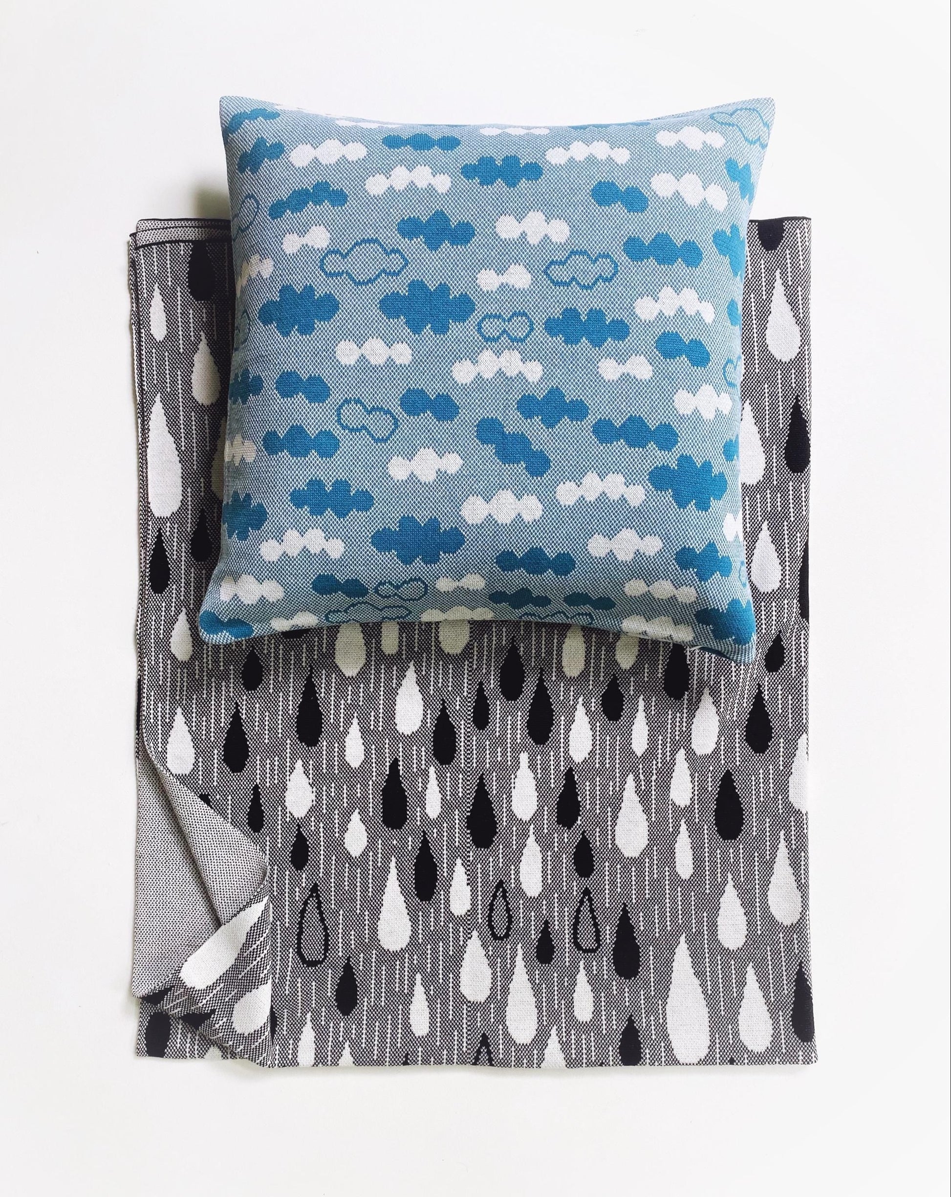 Blue decorative pillow with knitted cloud pattern on a folded knitted blanket with black and white raindrop design