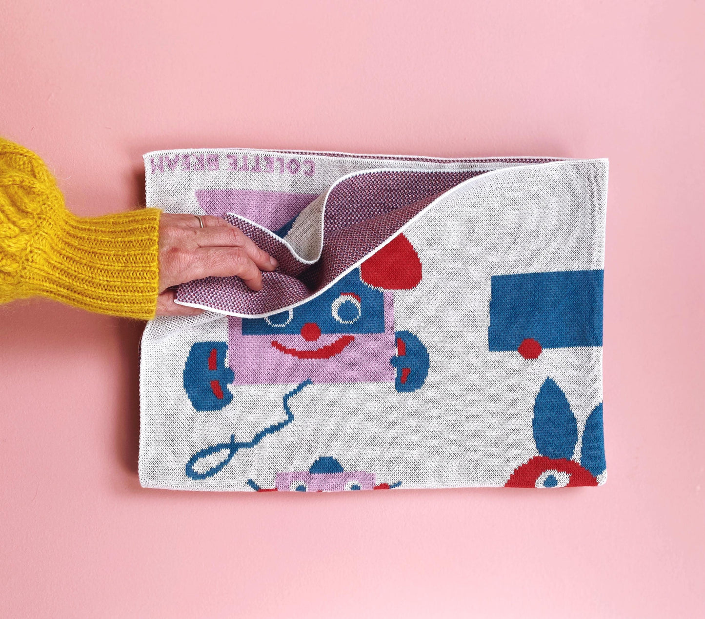 Person holding a colorful children's folded knit baby blanket with a cartoon design on a pink background