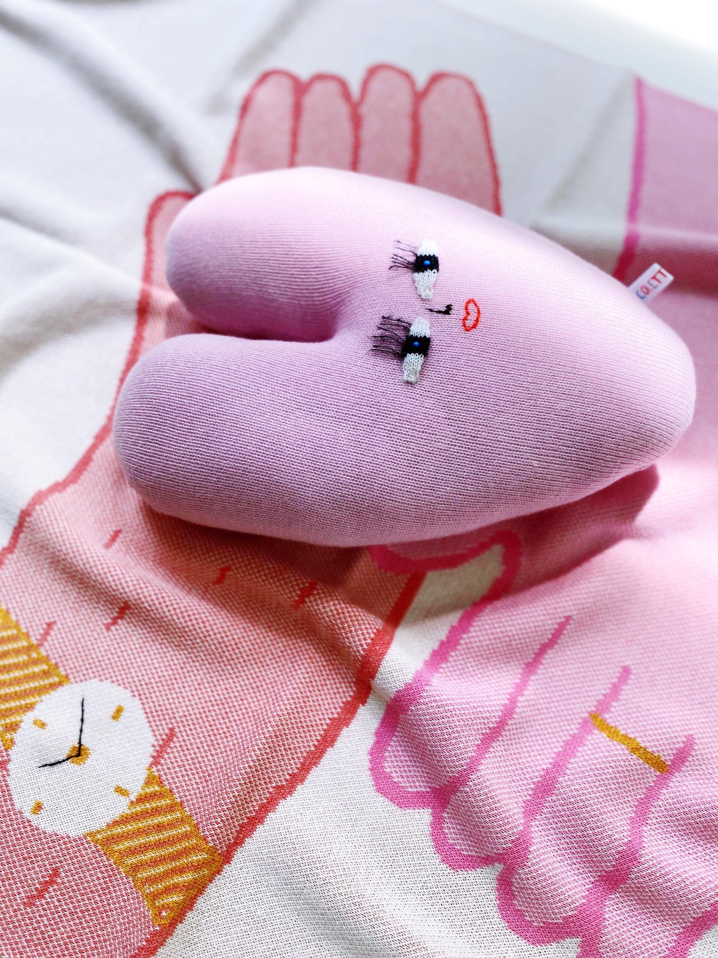 Pink plush toy resembling a heart with embroidered face details on a fabric background