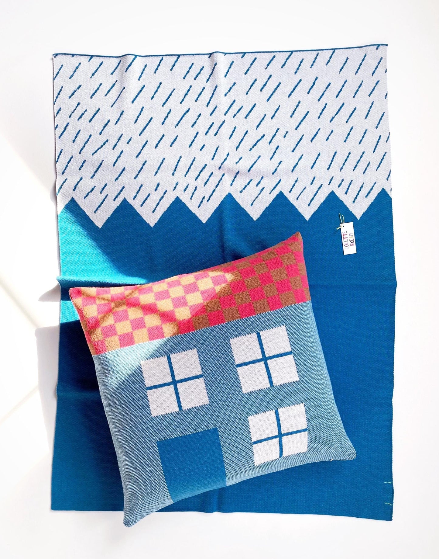 Blue knit blanket with geometric mountain design and a colorful house pillow on a white background