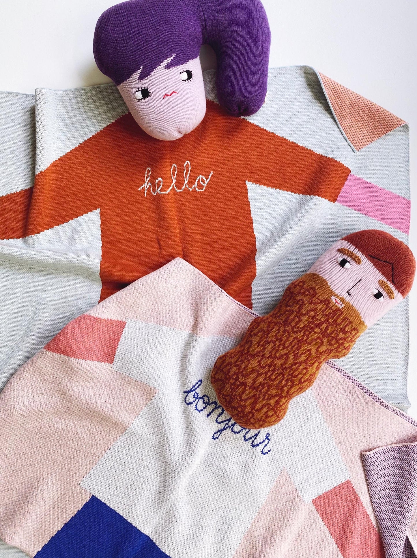 Two plush toys on a geometric-patterned blanket with 'hello' and 'bonjour' text.