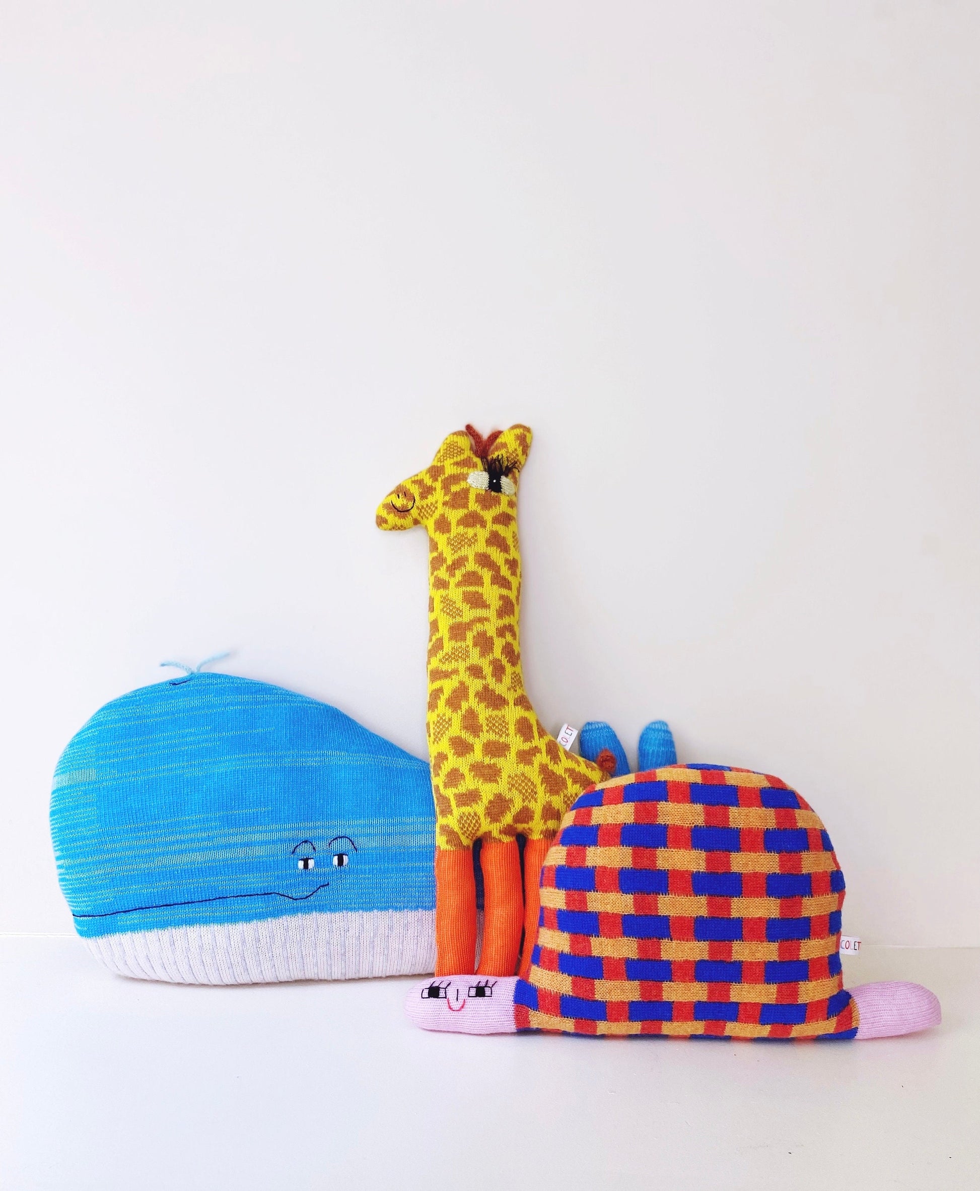 Colorful knitted lambswool plush toys including a giraffe, whale, and checkered turtle on a white background