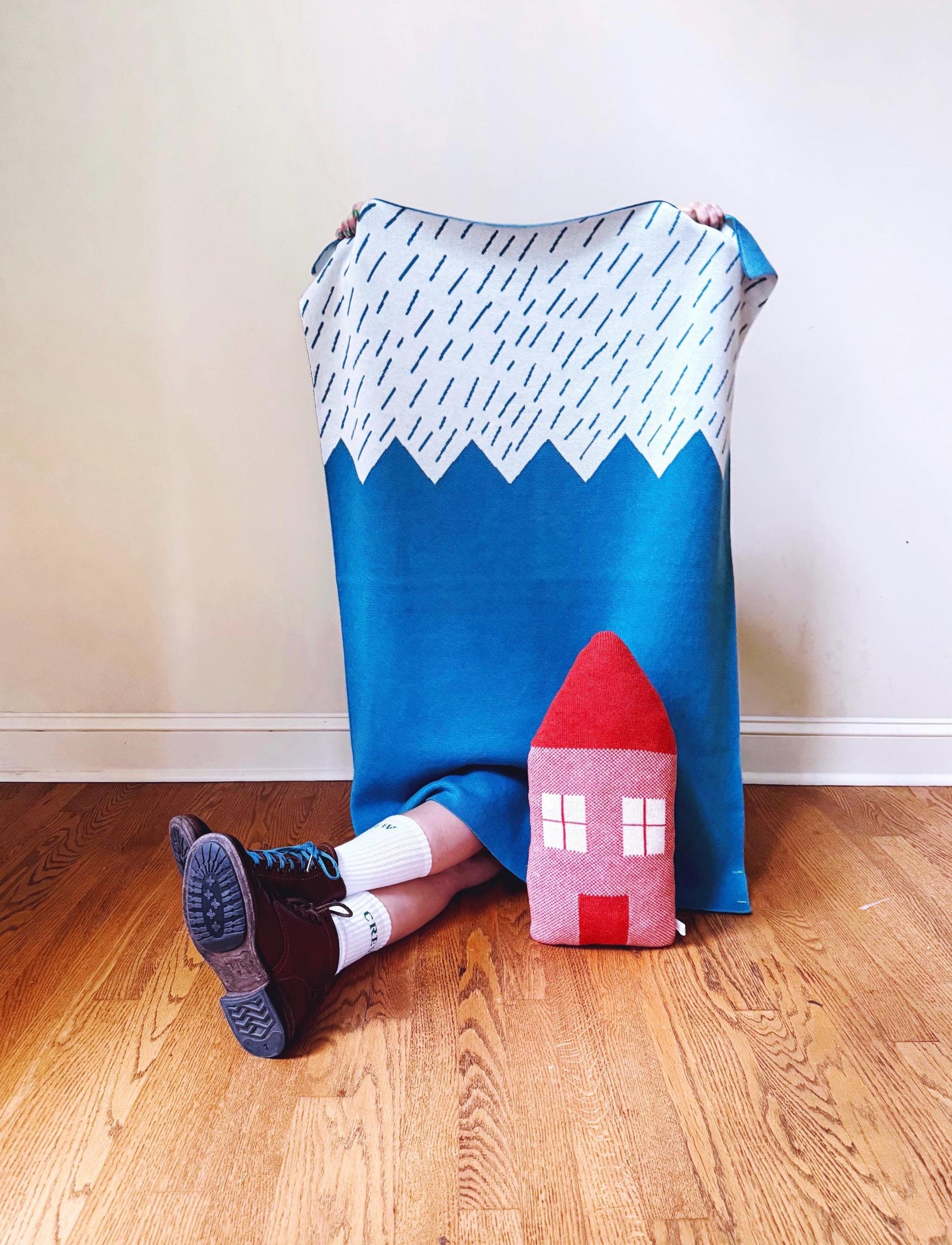 Person sitting on a wooden floor with a colorful blanket with a mountain design and a toy house shaped pillow.
